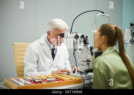 Optometrista anziano che esamina gli occhi del paziente utilizzando l'apparecchiatura in clinica. Paziente seduto sulla sedia mentre il medico esegue il controllo oculare Foto Stock