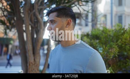 Il giovane ispanico in un abbigliamento informale cammina all'aperto in un vivace parco urbano con vegetazione, mostrando un comportamento calmo e un aspetto elegante ami Foto Stock