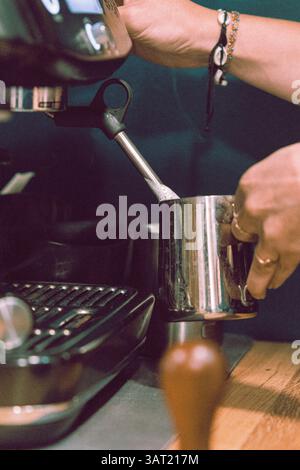 Primo piano di mani che montano il latte in una caraffa in acciaio inox utilizzando una macchina per espresso in un bar, preparandosi per la latte art. Foto Stock