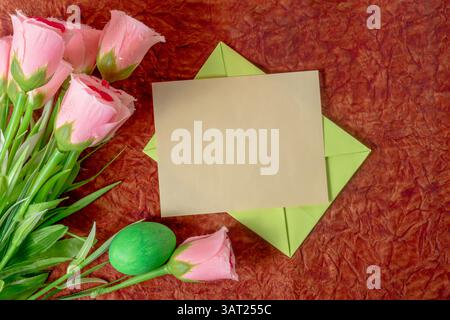 Un biglietto bianco in una busta verde circondata da rose rosa e un uovo di Pasqua verde, posto su una superficie rossa. Perfetto per Pasqua, biglietti d'auguri o Valen Foto Stock