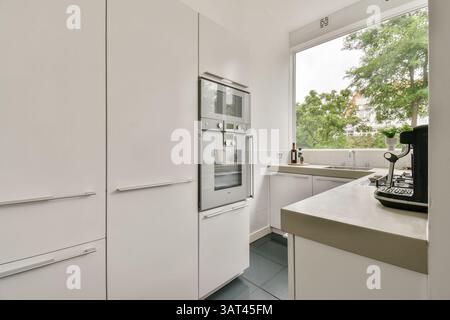 Cucina luminosa e spaziosa con eleganti mobili bianchi, elettrodomestici moderni e grandi finestre che rivelano il verde esterno, creando un ambiente di cottura sereno. Foto Stock