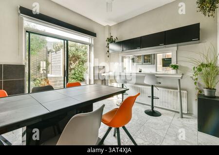 Elegante sala con tavolo nero e sedie arancioni vivaci, completata da luce naturale che filtra attraverso le grandi finestre, migliorando l'estetica moderna. Foto Stock