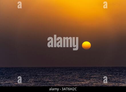 Un tramonto sereno sull'oceano, con un grande sole che tramonta all'orizzonte, proiettando un caldo bagliore dorato sul cielo e riflettendo sull'acqua. Foto Stock