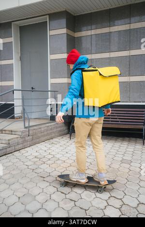 Corriere vestito con un abbigliamento casual in sella a uno skateboard, trasportando uno zaino giallo isolato pieno di cibo per una consegna tempestiva ai clienti in un ambiente urbano Foto Stock
