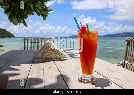 Bicchiere da cocktail con paglia blu, ciliegia e lime, su un tavolo di legno. Sullo sfondo il mare turchese Foto Stock