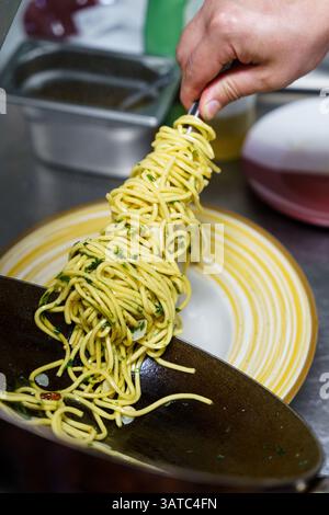 Uno chef prepara con cura una porzione di spaghetti aglio e olio utilizzando pinze in una cucina professionale. La pasta è ricoperta di olio d'oliva, aglio, prezzemolo, Foto Stock