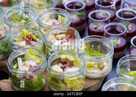 Servizio catering per preparare insalata caesar fresca con lattuga, crostini, pancetta e parmigiano in vasetti di vetro per l'evento Foto Stock