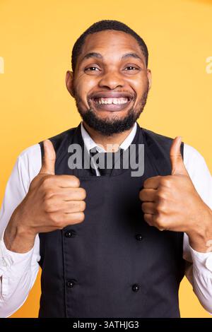 Maggiordomo felice e sicuro che dà un simbolo di pollice davanti alla telecamera, presentando il suo accordo e le emozioni positive in studio. Giovane cameriere che mostra un segno simile. Foto Stock