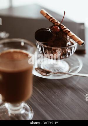Tazza di caffè con latte, gelato al cioccolato con sciroppo dolce, waffle, biscotti e ciliegie fresche, delizioso dessert in una ciotola di vetro Foto Stock