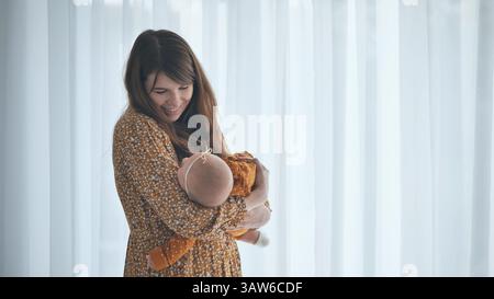 Madre felice che tiene in braccio la bambina appena nata addormentata, in piedi vicino alla finestra con tende bianche Foto Stock
