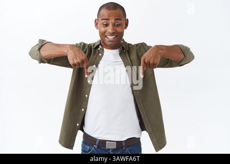 Ritratto di uomo felice allegro, eccitato, maschio afroamericano che punta verso il basso, vedendo buona offerta, sconto speciale, finalmente trovato buon affare, in piedi w Foto Stock
