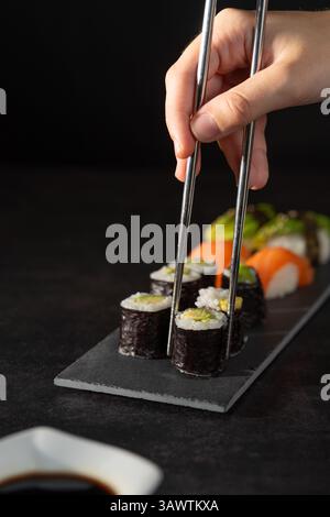 Una mano che prende un pezzo di maki di avocado con bacchette in acciaio inox da un piatto di sushi. Contiene più makis, nigiri di salmone e nigiri di avocado. TH Foto Stock