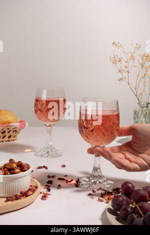 Due bicchieri di vino rosa su una tovaglia bianca, una mano che prende uno di loro. Sul tavolo c'è una piccola ciotola con arachidi e uva rossa. Foto Stock