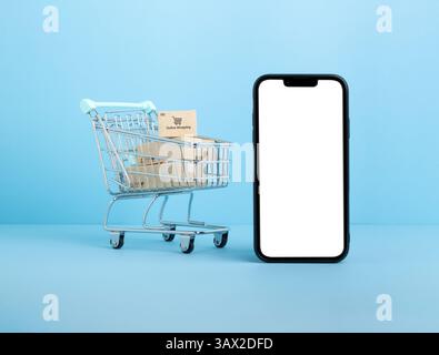 Carrello pieno di piccoli pacchi posizionati accanto a uno smartphone con una schermata bianca vuota su sfondo blu, che rappresenta lo shopping online, e-co Foto Stock