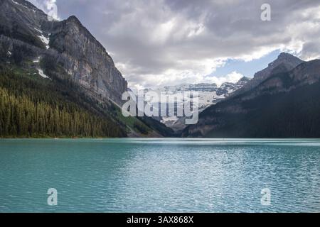 L'acqua turchese riflette le vette frastagliate e la vegetazione lussureggiante che circonda un tranquillo lago. Le nuvole si spostano sopra la testa, creando un tranquillo e pittoresco atmo Foto Stock
