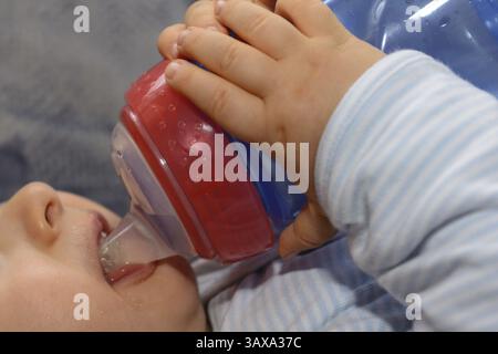 Il bambino beve il tè da un biberon e tiene il biberon con entrambe le mani - primo piano Foto Stock
