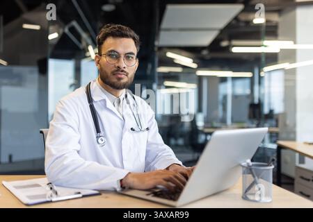 Un medico sicuro si siede alla sua scrivania e lavora su un notebook in un ufficio moderno. Indossa uno stetoscopio e guarda la telecamera. Foto Stock