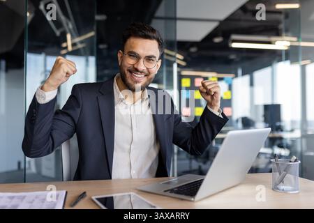 Un giovane uomo d'affari di successo festeggia, alzando i pugni in aria con un ampio sorriso alla scrivania dell'ufficio con un notebook. Foto Stock