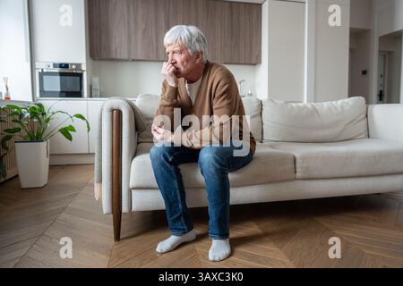 Un pensionato malato con demenza che copre la bocca, seduto sul divano in difficoltà. Paura per la perdita di memoria Foto Stock