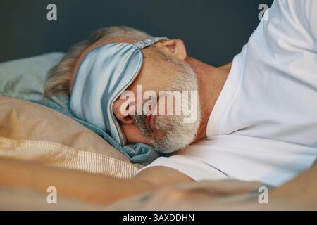 Uomo anziano che indossa una maschera dormiente mentre riposa tranquillamente sul letto con cuscino e coperta, catturando un momento sereno di relax Foto Stock