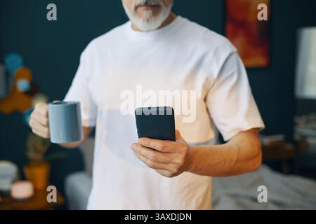 Uomo anziano che tiene in mano smartphone e tazza mentre si trova in salotto. Concentrati sul dispositivo e goditi momenti indimenticabili in un ambiente domestico confortevole Foto Stock