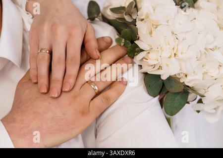 Una vista ravvicinata delle mani delle coppie di sposati si sono unite, mostrando le loro fedi nuziali d'oro, con un lussureggiante bouquet bianco accanto ad esse. Foto Stock