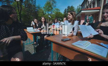 Adolescenti allegri che studiano e chiacchierano insieme alle scrivanie all'aperto in una giornata di scuola soleggiata Foto Stock