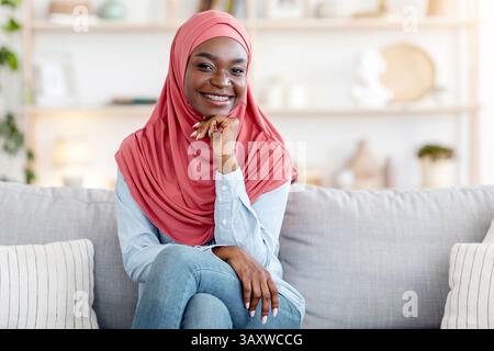 Gioiosa giovane musulmana africana in hijab in posa sul divano a casa Foto Stock