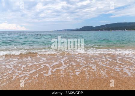 Tranquilla spiaggia caratterizzata da onde morbide che si infrangono sulle coste sabbiose con colline in lontananza sotto un cielo parzialmente nuvoloso. Perfetto per concetti di relax Foto Stock