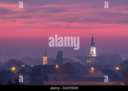 Tranquilla vista serale di un paesaggio urbano caratterizzato da torri e case della chiesa adagiate su un vibrante cielo al tramonto, evocando sentimenti di serenità e pace Foto Stock