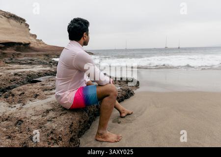 Un uomo seduto su una roccia in spiaggia che guarda l'oceano e si rilassa. Foto Stock
