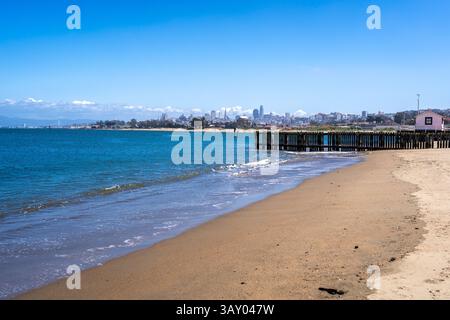 La spiaggia sabbiosa di Golden Gate Beach incontra l'Oceano Pacifico, con lo skyline di San Francisco visibile in lontananza sotto un cielo azzurro Foto Stock