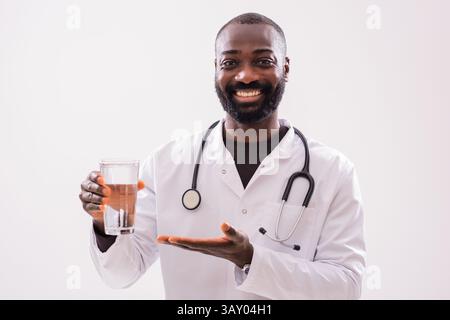 Ritratto di allegro medico africano in uniforme medica che sta mostrando un bicchiere d'acqua su sfondo grigio. Foto Stock