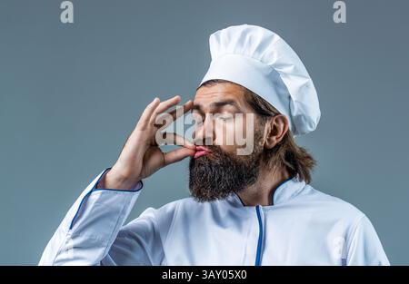 Lo chef fornello in grembiule mostra segni di buon gusto, gesto bellissimo, bacio dita sfondo grigio isolato. Ritratto di un cuoco felice. Cappello da cucina Foto Stock
