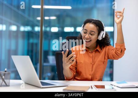Una donna entusiasta in ufficio, che indossa cuffie e occhiali, è entusiasta mentre guarda il suo telefono. La scena include un notebook e un blocco note. Foto Stock