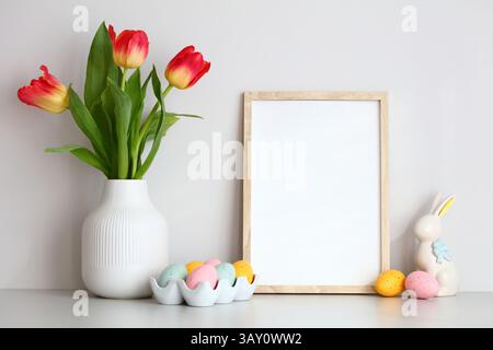 Buona Pasqua, arredamento su tavolo su sfondo bianco. Modello cornice, vaso di tulipani rossi, uova di Pasqua e coniglietto. Foto Stock