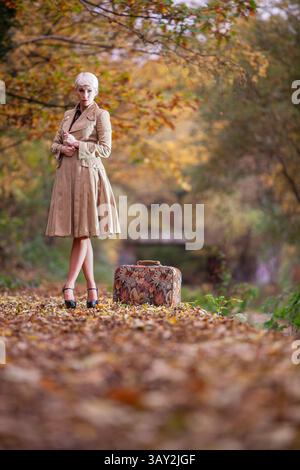 Una bella giovane donna che aspetta su una piattaforma rurale e umnale con una valigia e un abbigliamento retrò. Foto Stock