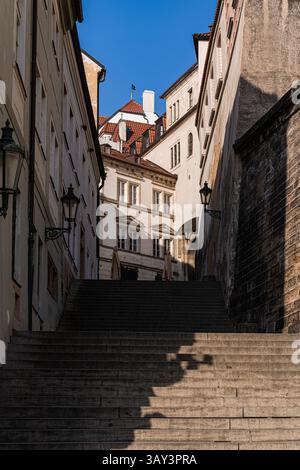 Una stretta strada medievale con ripide scale in pietra, circondata da edifici storici con finestre ornate e affascinanti lanterne a Praga. Sole brillante Foto Stock