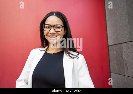 Una donna ispanica sicura di sé che indossa occhiali e abbigliamento da lavoro si staglia su uno sfondo rosso e grigio, trasudando professionalità ed eleganza in un moderno Foto Stock