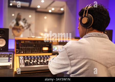 Un produttore musicale che indossa le cuffie, concentrato sul mixaggio audio in uno studio di registrazione professionale. Un musicista suona la chitarra dietro una finestra di vetro, creato Foto Stock