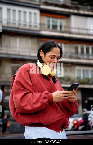 Un uomo con capelli lunghi e grandi occhiali con una giacca rossa e cuffie gialle è fuori a guardare uno smartphone, con un edificio urbano nella ba Foto Stock
