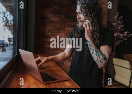 Un uomo barbuto con lunghi capelli e tatuaggi usa un computer portatile mentre parla al telefono, posizionato vicino a una grande finestra in un ambiente accogliente. Ligh naturale Foto Stock