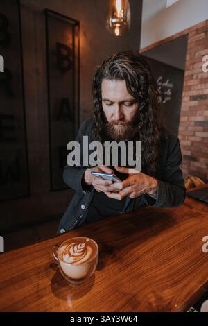 Un uomo barbuto usa intensamente il suo smartphone mentre è seduto ad un bancone di legno in un accogliente bar. Una tazza di caffè con latte art si trova vicino al bancone Foto Stock