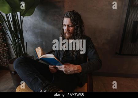 Un uomo con i capelli lunghi e la barba sta leggendo un libro seduto in una comoda poltrona di un caffè. L'ambiente è accogliente, con grandi piante in vaso e. Foto Stock