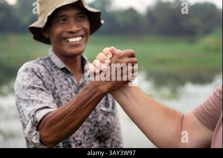 Primo piano di un contadino indonesiano sorridente che stringe la mano a un'altra persona, incarnando fiducia e cooperazione. Sullo sfondo sono presenti terreni agricoli lussureggianti e verdi a. Foto Stock