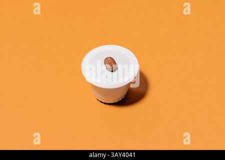 Una capsula di caffè biodegradabile posizionata su un vivace sfondo arancione, sormontata da un unico chicco di caffè, che mostra semplicità e modernità Foto Stock