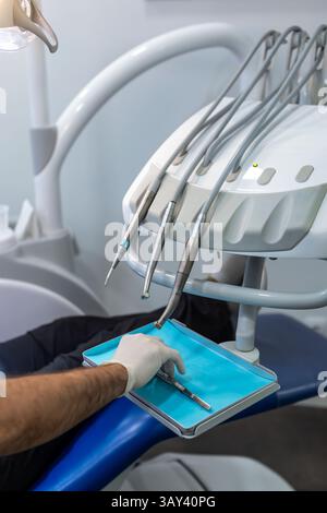 Primo piano di una mano indossata dai guanti da dentista che cerca uno strumento in mezzo a attrezzature dentali avanzate. L'immagine cattura l'essenza di un odontoiatrico contemporaneo Foto Stock