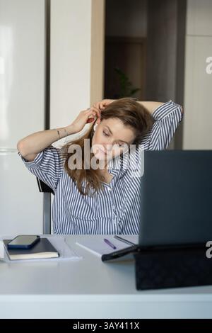 Donna con una camicia a righe a casa, che regola le cuffie mentre lavora su un laptop. Un telefono, un notebook e una penna sono sul tavolo, creando un lavoro accogliente Foto Stock