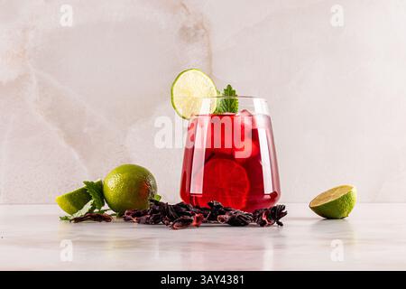 Un bicchiere di rinfrescante tè rosso all'ibisco con cubetti di ghiaccio, guarnito con una fetta di lime e foglie di menta. Circondati da ingredienti freschi e secchi, pronti Foto Stock