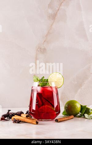 Un vivace tè freddo all'ibisco guarnito con lime e menta in vetro su una superficie in marmo. Circondato da bastoncini di cannella e lime, crea una rinfrescante sensazione Foto Stock
